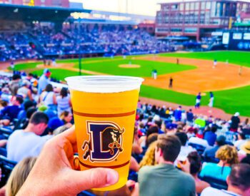 close up of a beer with a crowd of people watching a baseball game in the background