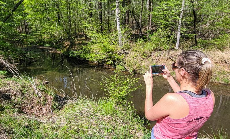 A person taking a picture of a river, standing on a hiking trail