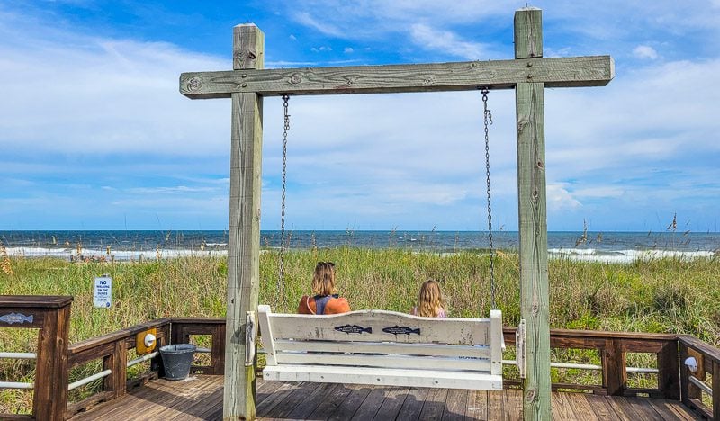 A mother and child sitting on a wooden swing on Carolina Beach boardwalk with views of the ocean