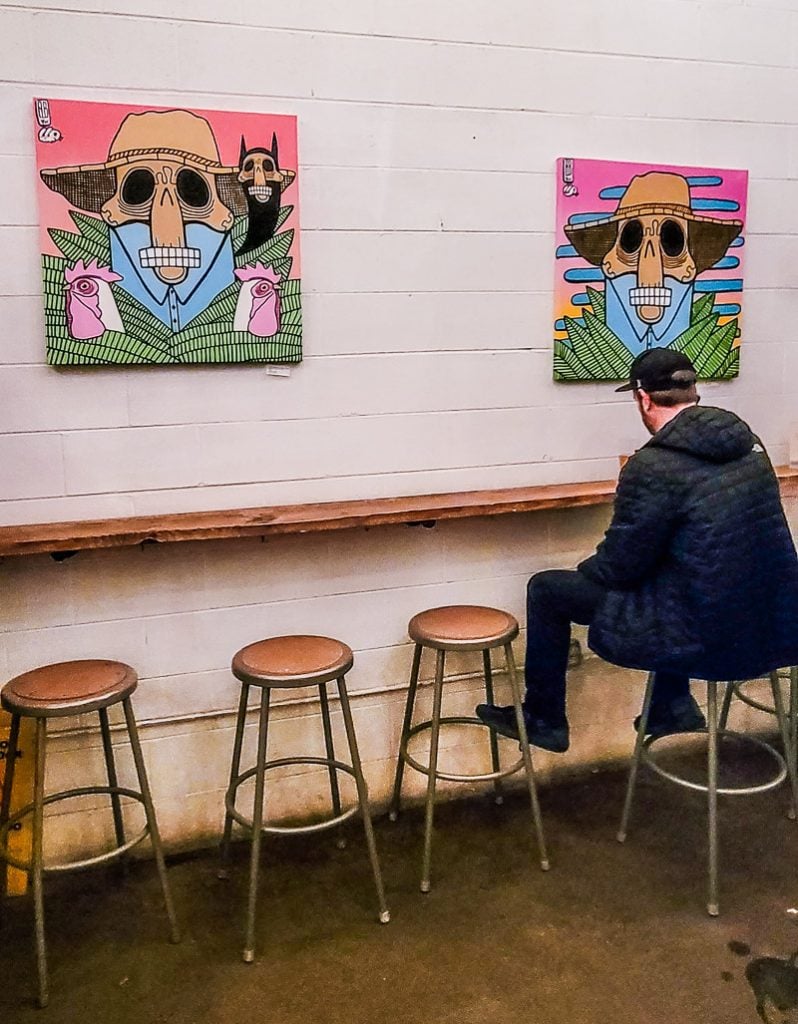 Man sitting on a stool drinking beer with two pieces of art work.
