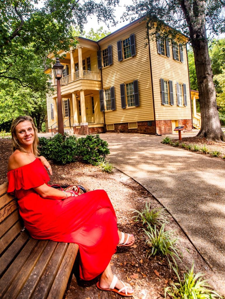 Lady in red dress sitting on bench with historic home behind her