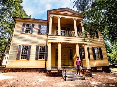Man in a pink shirt standing on steps in front of a historic yellow house