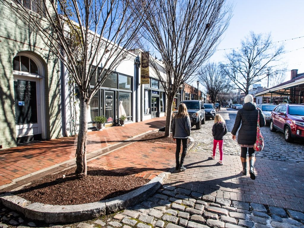 Three people walking down a cobblestone street with shops on the sidewalk.