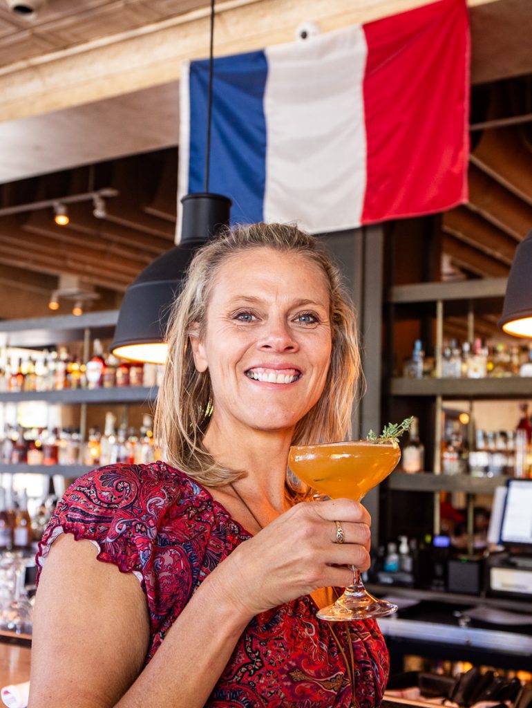 Woman drinking a cocktail at a bar under a French flag.