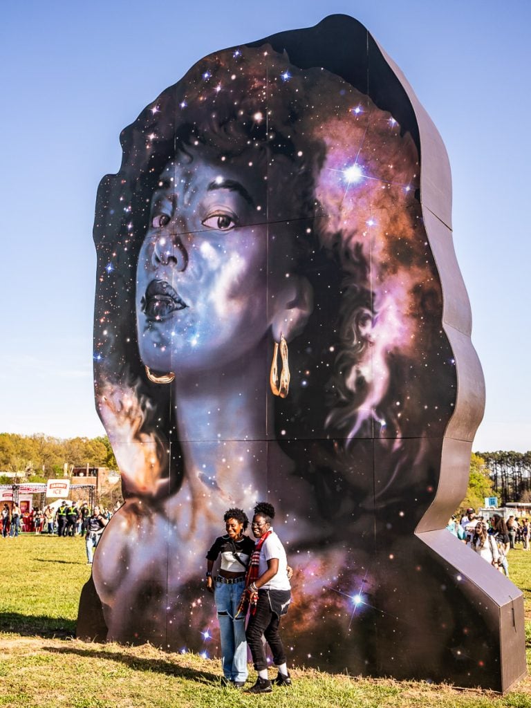 People at a music festival in front of a monument of a woman.