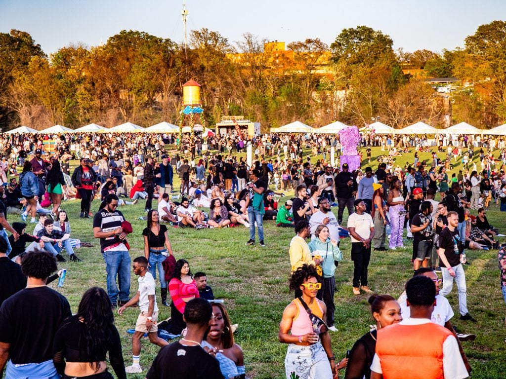 People at a music festival walking and sitting on a grass field.