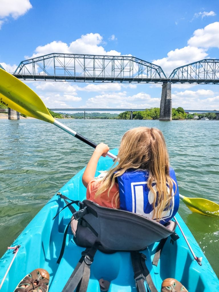 Girl kayaking down a river with a bridge in the distance.
