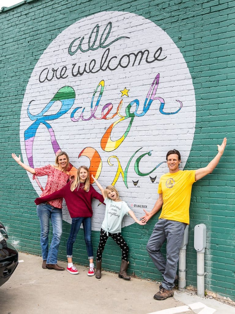 Family of four posing in front of a mural that says all are welcome to Raleigh.