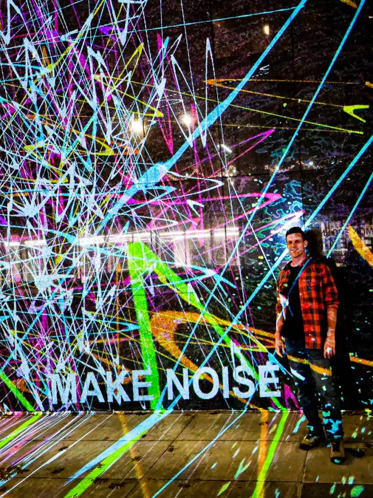 man standing in front of paint spllatered light show with make some noise sign