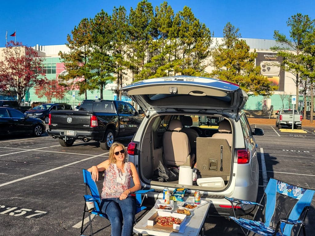 A lady sitting in camp chairs tailgating in a car park at a sports arena.