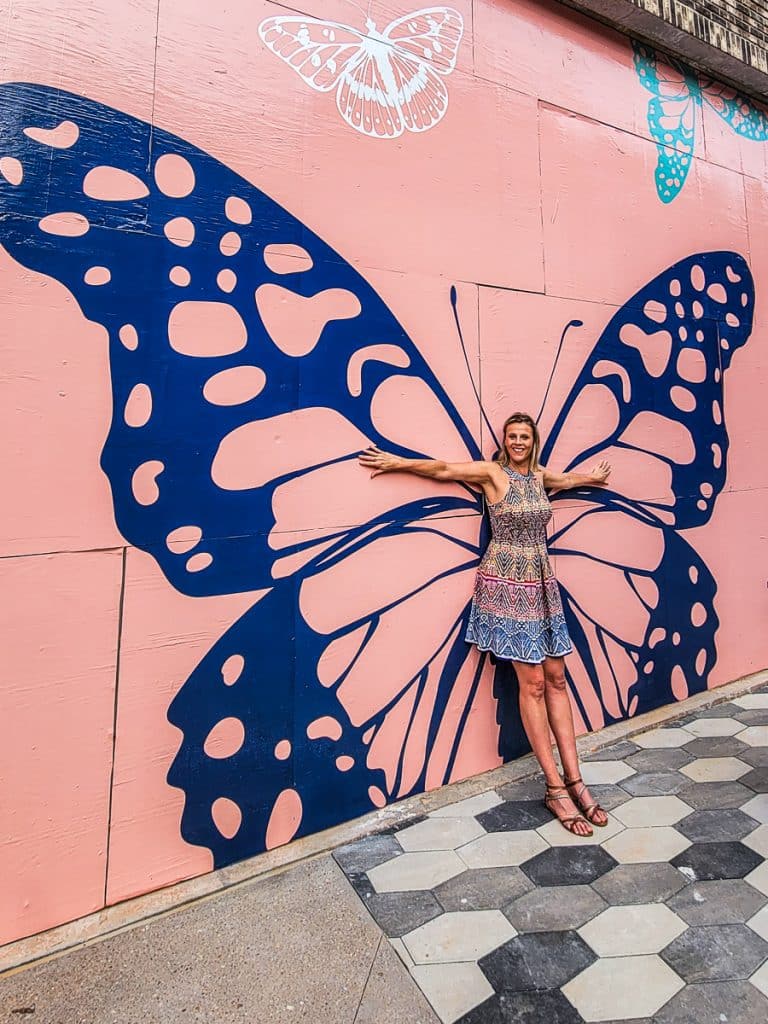 Lady standing against a mural of a butterfly on a wall.