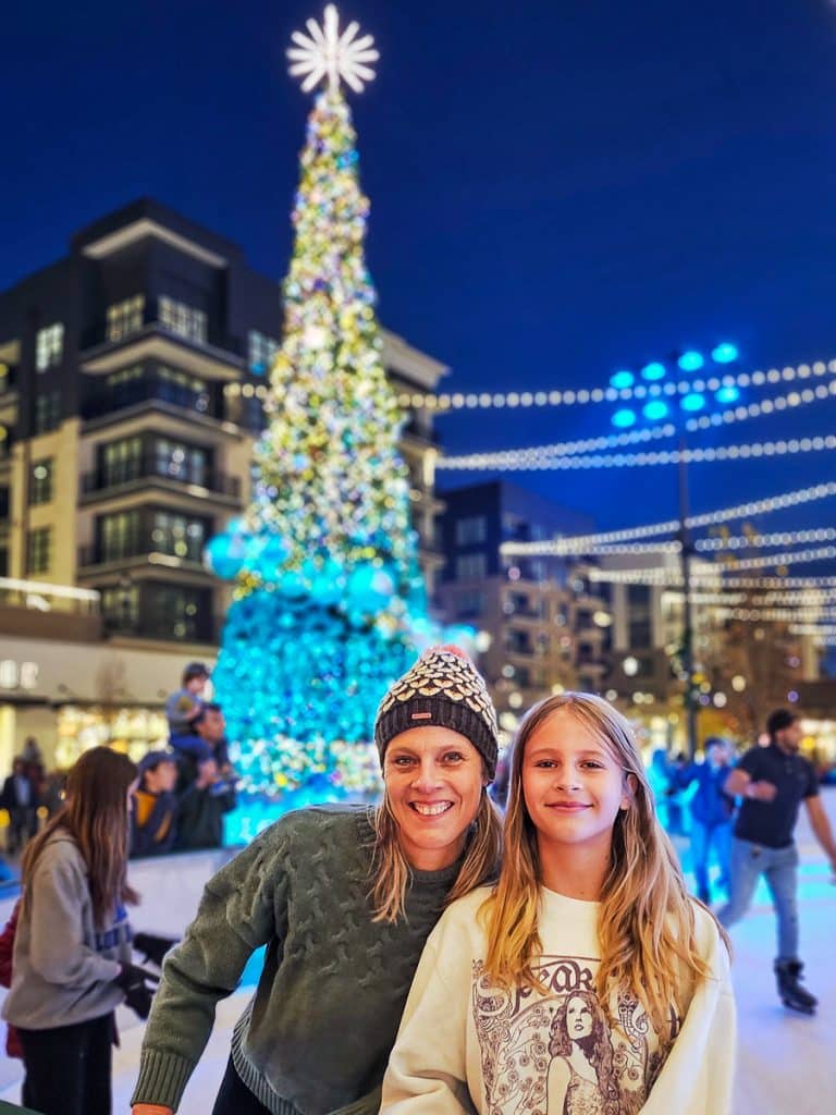 Mom and daughter ice skating in front of a Christmas tree.