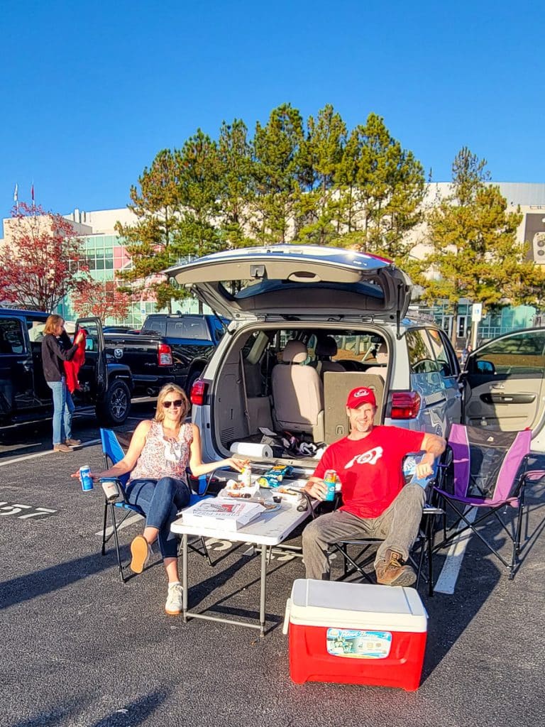 Man and woman sitting in camp chairs tailgating outside a sports arena.