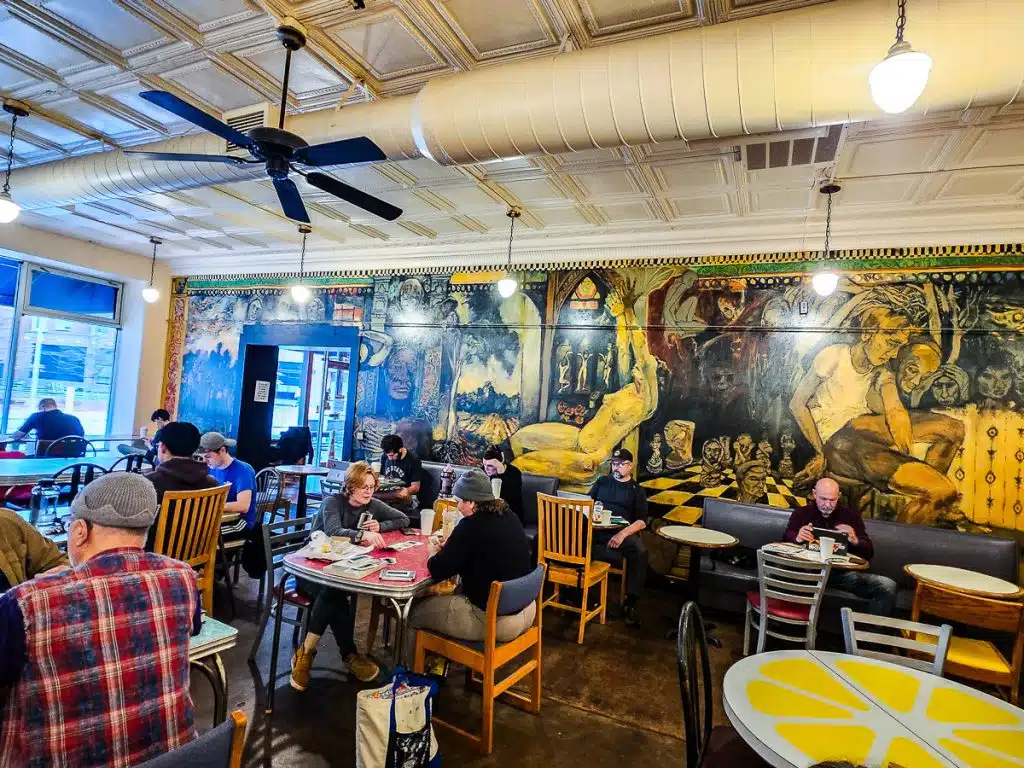 People sitting in a coffee shop at tables and chairs with a mural on the wall.