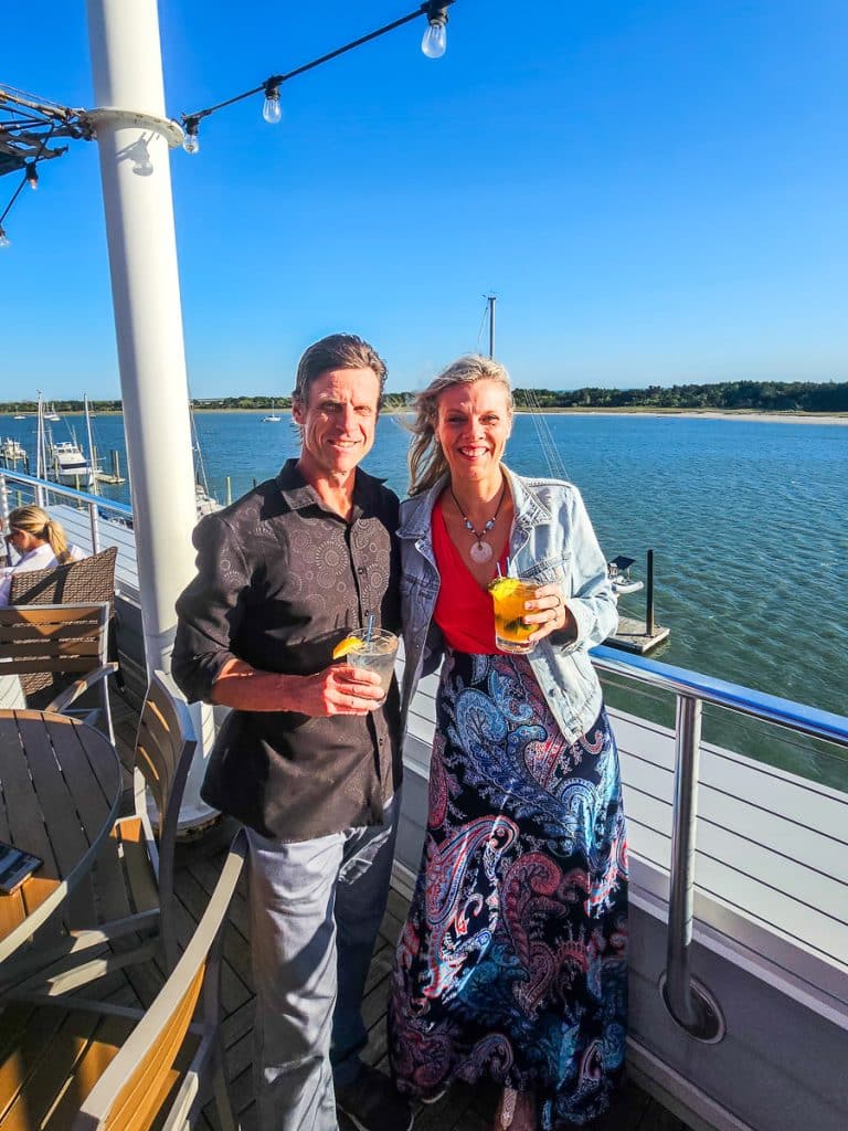 A couple drinking cocktails standing up with water views.