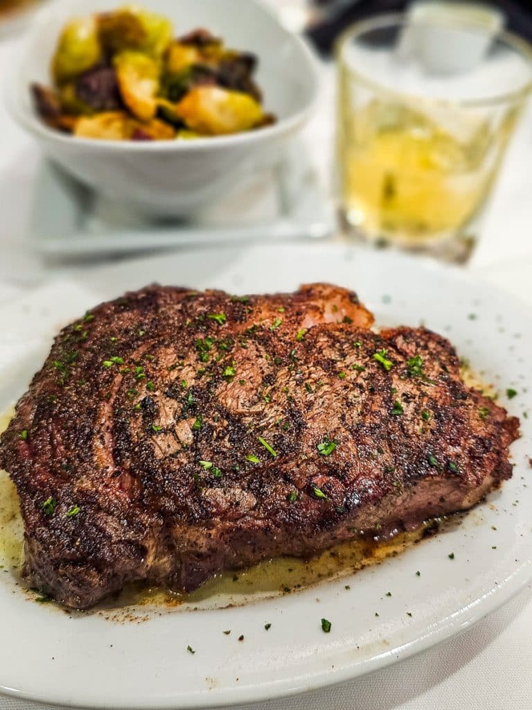 Steak on a plate from Ruth's Chris with Brussel sprouts and a cocktail in the background