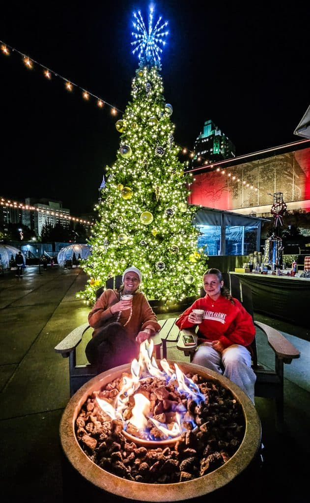 mother and daughter drinking hot chocolate by firepit and christmas tree at the rink